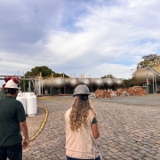 Três pessoas utilizando capacetes de proteção caminham a céu aberto, em vistoria da Fepam. Ao fundo, botijões e grandes tanques de combustível