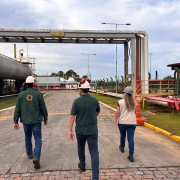 Três pessoas utilizando capacetes de proteção caminham a céu aberto em complexo industrial