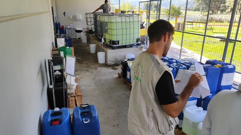 Duas pessoas realizam an&aacute;lises qu&iacute;micas. No local, diversos gal&otilde;es com produtos