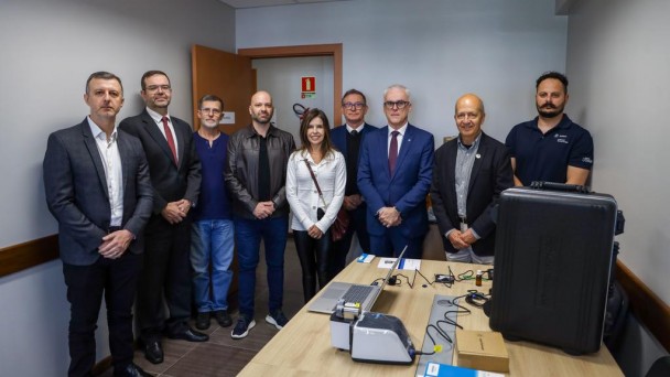 No primeiro plano, mais &agrave; direita, nateriais de laborat&oacute;rio aparecem dispostos sobre mesa de madeira. No fundo, nove pessoas, uma ao lado da outra